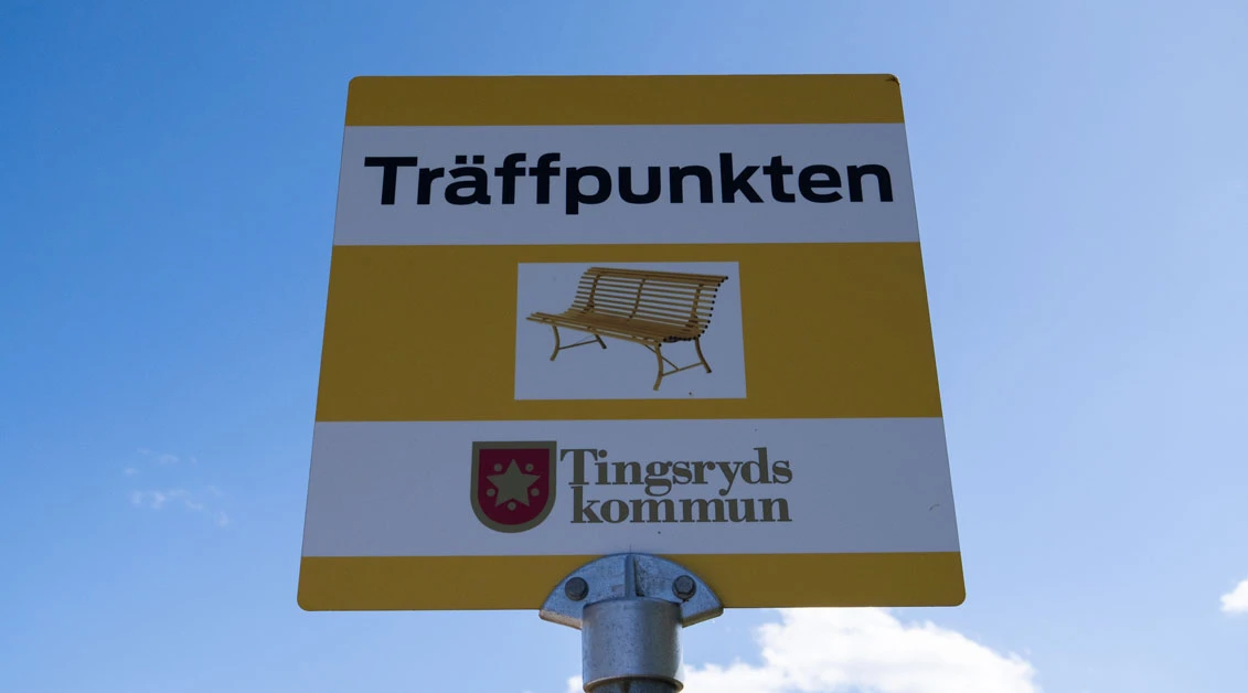 Skylt med texten "Träffpunkten" och en symbol av en bänk samt Tingsryds kommuns logotyp, mot en klar blå himmel.