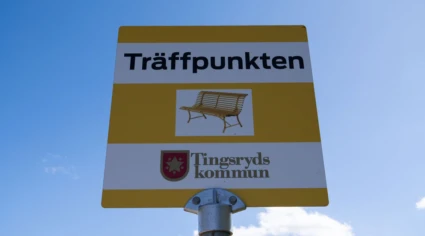 Skylt med texten "Träffpunkten" och en symbol av en bänk samt Tingsryds kommuns logotyp, mot en klar blå himmel.