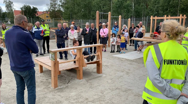 En grupp människor samlade vid ett utegym i en park. En person visar en träningsbänk medan andra tittar på. Några funktionärer i gula reflexvästar med texten "FUNKTIONÄR" övervakar evenemanget. I bakgrunden syns fler träningsredskap, ett stängsel och grönska.