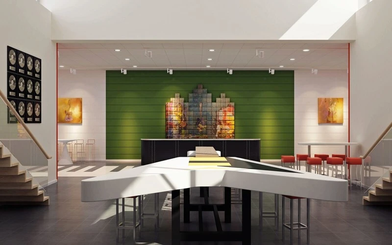 Ett modernt inrett rum med ett stort vitt bord i centrum format som en pil, omgiven av höga stolar. Rummet har en grön fondvägg med en färgstark konstinstallation och är upplyst av takfönster som släpper in naturligt ljus. På väggarna hänger tavlor och till vänster syns guldskivor uppsatta i ram.