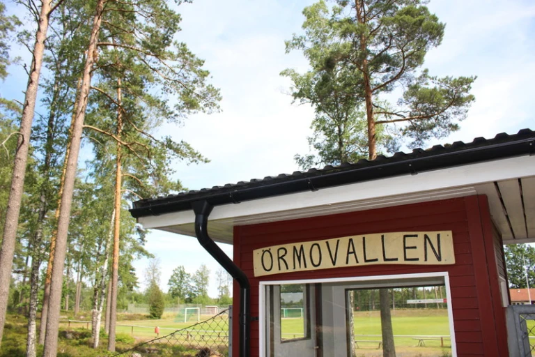 En skylt med texten "ÖRMOVALLEN" är monterad på en röd byggnad med vit takfot. I bakgrunden syns en fotbollsplan omgiven av höga träd och ett staket.