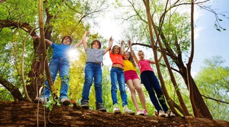 Fem barn som står på en stor trädstam i en skog. De håller varandra i händerna och ser ut att ha roligt tillsammans. Solen skiner genom träden och skapar en ljus och glad atmosfär.