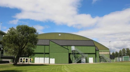 Ishallen har ett välvt tak. Det finns en metalltrappa på sidan av byggnaden och ett träd till vänster. Himlen är delvis molnig och det finns en välskött gräsyta i förgrunden.