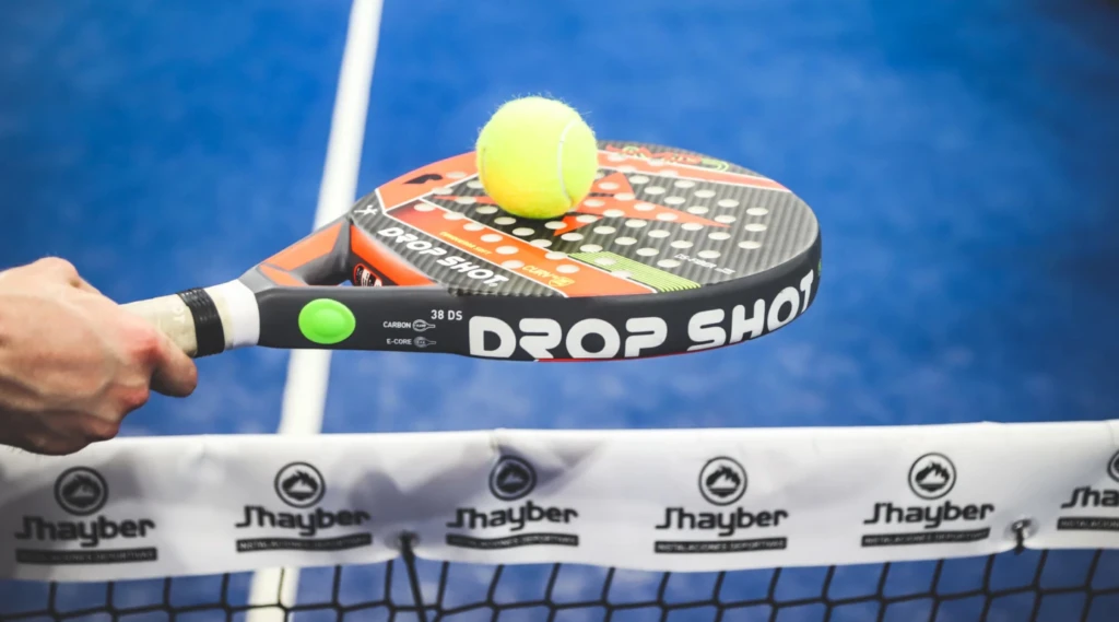 En närbild av ett padelracket som träffar en gul tennisboll över ett nät.