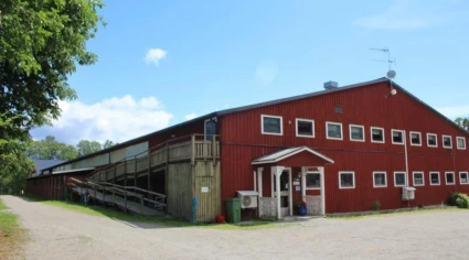 Bilden visar en stor röd laduliknande byggnad med flera fönster och en liten ingångsveranda. Det finns en ramp på vänster sida av byggnaden som leder till en övre nivå. Byggnaden är omgiven av träd och har en klarblå himmel ovanför sig.