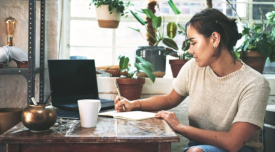 En person sitter vid ett träbord i ett rum fyllt med växter. Personen skriver i en anteckningsbok med en penna