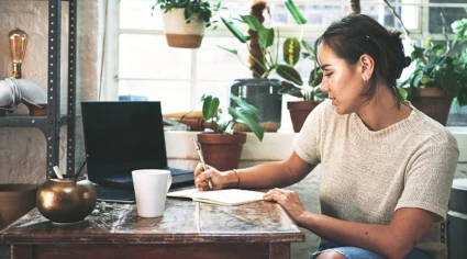 En person sitter vid ett träbord i ett rum fyllt med växter. Personen skriver i en anteckningsbok med en penna