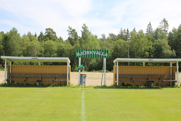 Bilden visar en fotbollsplan med två avbytarbänkar, en på varje sida av planen. Ovanför bänkarna finns skyltar som markerar "HEMMA" och "BORTA". I mitten syns en skylt med texten "BJÖRKVALLA". Bakom planen finns träd och skog.