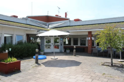 Utegård med stenlagd mark, vit parasoll, blå uppblåsbar pool och krukväxter. I bakgrunden syns en byggnad med stora fönster och grönt tak med växtlighet samt bord och stolar under tak.