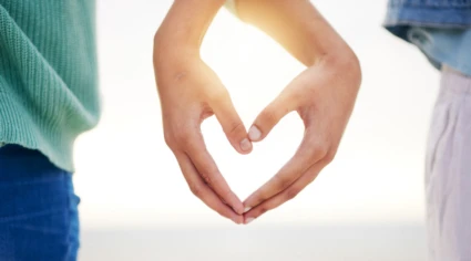 Två personer formar ett hjärta med sina händer mot en ljus bakgrund. Personen till vänster bär en grön tröja och blå jeans, medan personen till höger har en ljus topp och byxor.