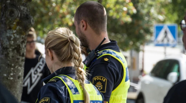 Två poliser med uniformer märkta 'POLIS' står utomhus. Den ena har flätad frisyr. I bakgrunden syns en person med suddat ansikte, ett träd, parkerade bilar och en skylt för övergångsställe.