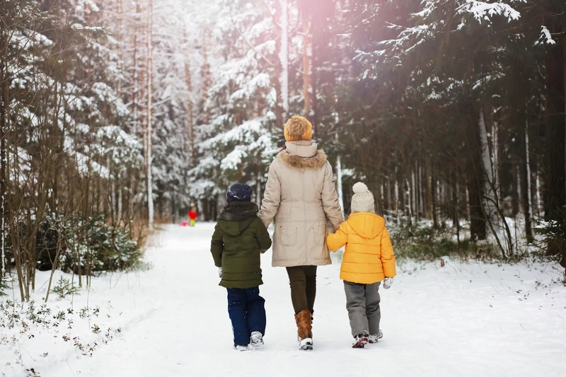 Tre personer går på en snötäckt stig i en skog. Den mittersta personen, iklädd beige jacka med pälskantad huva och gul mössa, håller barnens händer. Barnet till vänster har grön jacka och blå byxor, barnet till höger har orange jacka och grå byxor. Snötäckta träd omger dem, och längre fram syns ytterligare en person i röd jacka.