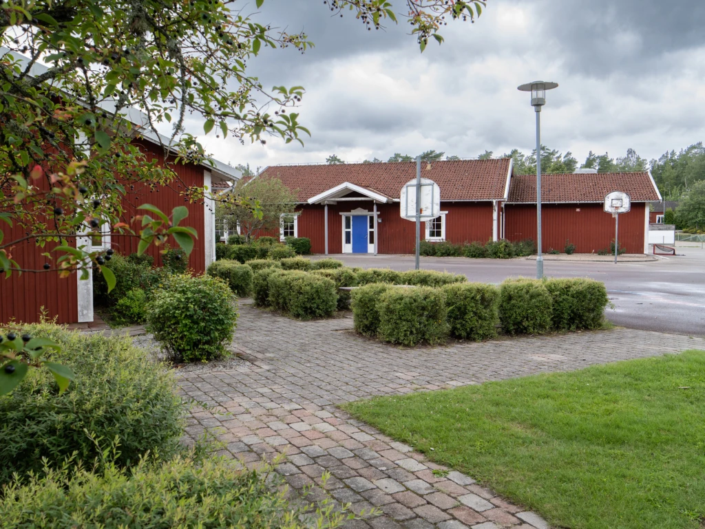 Röda skolbyggnader med utegård med gräs, asfalt, buskar och basketkorg i förgrunden