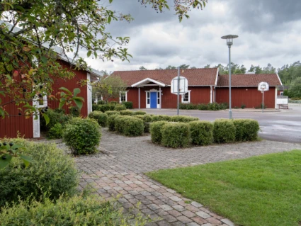 Röda skolbyggnader med utegård med gräs, asfalt, buskar och basketkorg i förgrunden