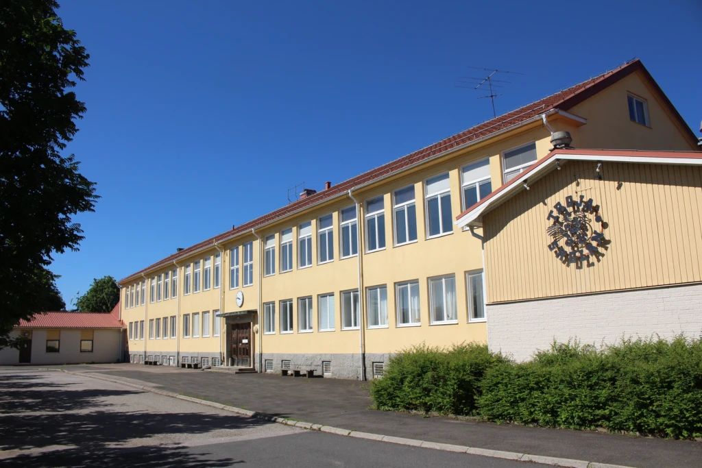 Gul skolbyggnad omringad av asfalt, buskar och träd.