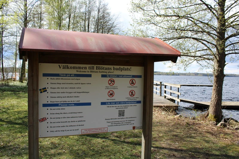 Informationsskylt vid Blötans badplats, omgiven av träd och med en brygga som sträcker sig ut i sjön i bakgrunden. Skylten innehåller regler och tips på svenska och engelska, samt symboler för förbjudna aktiviteter som hundar, rökning, alkohol och eldning. En QR-kod finns för mer information.