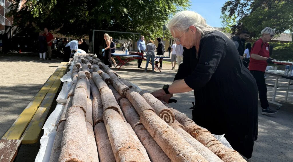 Långa rader av rulltårtor ligger uppdukade på ett bord utomhus under ett evenemang, med människor i bakgrunden som förbereder och ordnar servering.