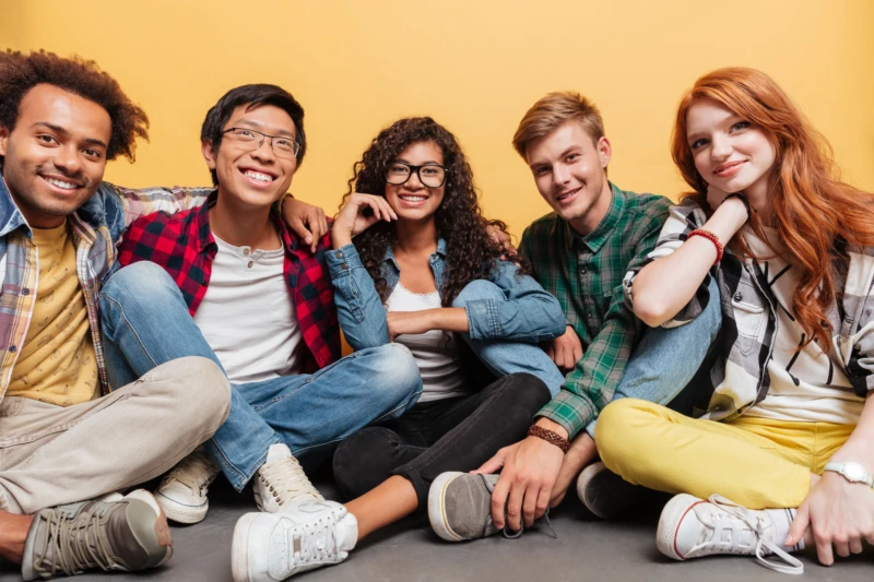 En grupp unga personer sitter avslappnat p&aring; golvet framf&ouml;r en gul bakgrund, kl&auml;dda i f&auml;rgglada och vardagliga kl&auml;der.