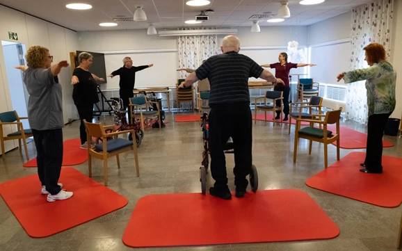 En grupp seniorer deltar i balansträning i ett rum med stolar och röda träningsmattor på golvet. Deltagarna står utspridda och gör armrörelser under ledning av en instruktör längst fram. I mitten står en deltagare med rollator. Rummet är ljust med stora fönster och gardiner, och stolar är placerade runt övningsytan.