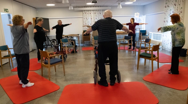 En grupp seniorer deltar i balanstr&auml;ning i ett rum med stolar och r&ouml;da tr&auml;ningsmattor p&aring; golvet. Deltagarna st&aring;r utspridda och g&ouml;r armr&ouml;relser under ledning av en instrukt&ouml;r l&auml;ngst fram. I mitten st&aring;r en deltagare med rollator. Rummet &auml;r ljust med stora f&ouml;nster och gardiner, och stolar &auml;r placerade runt &ouml;vningsytan.