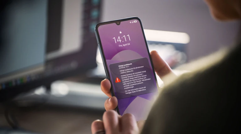 Person håller en mobiltelefon i handen inomhus och tittar på ett viktigt myndighetsmeddelande (VMA) på skärmen, med varning om en händelse i Malmö och uppmaning att stanna inomhus och följa information.