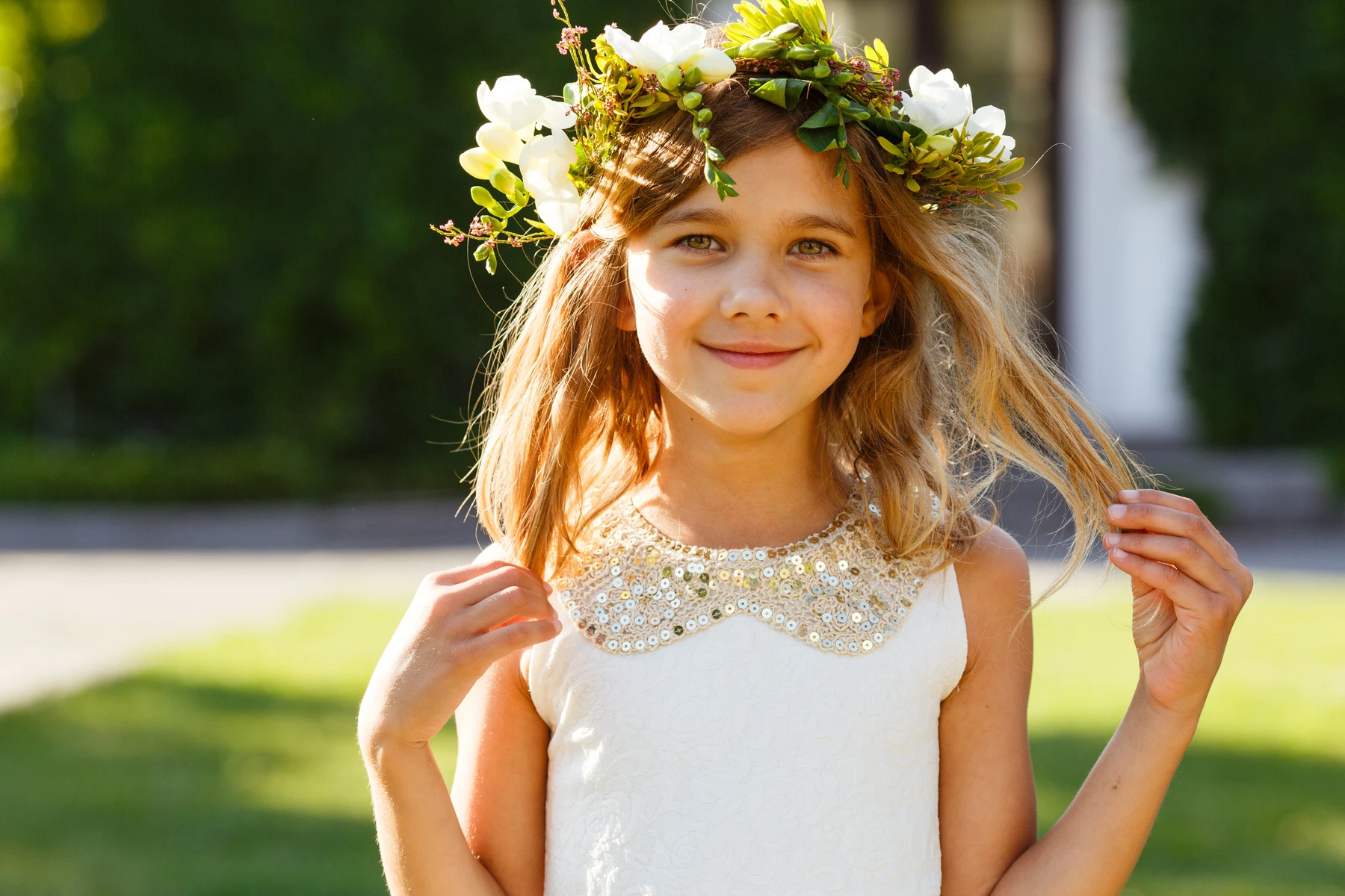 Barn med ljus sommarklänning och blomsterkrans på huvudet står utomhus i soligt väder och håller i sitt hår. I bakgrunden syns grönska och mjukt suddig naturmiljö.